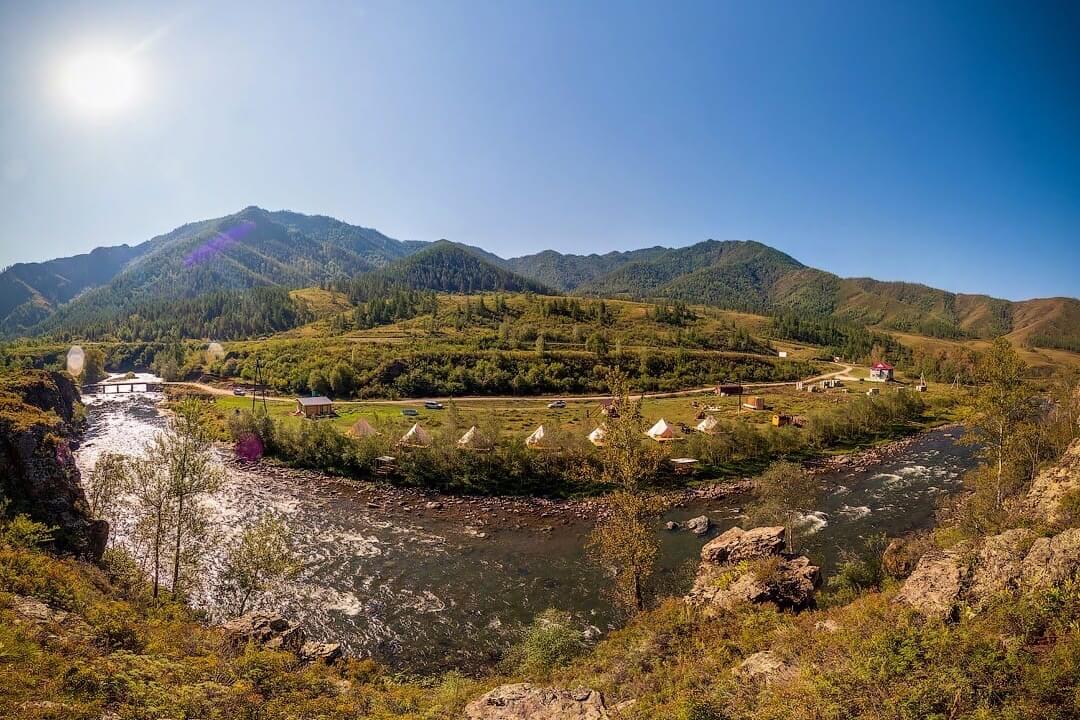 Панорамный снимок: горы и солнце, бурное течение горной реки и территория глэмпинга.