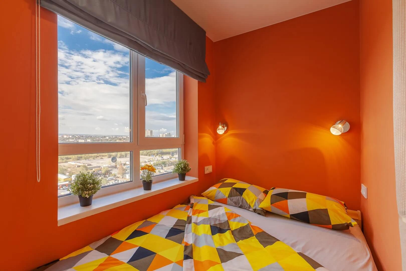 Кровать расположена у окна. Стены выкрашены в яркий оранжевый цвет.