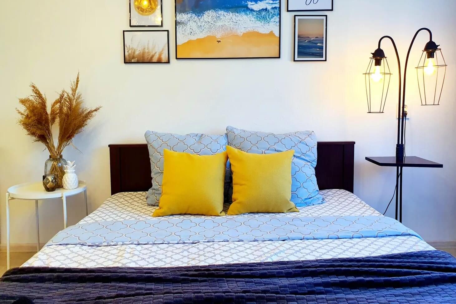 Интерьер спальни: ваза с сухостоями, картины над изголовьем кровати и оригинальной формы светильник. В центре кровати - две яркие желтые подушки.