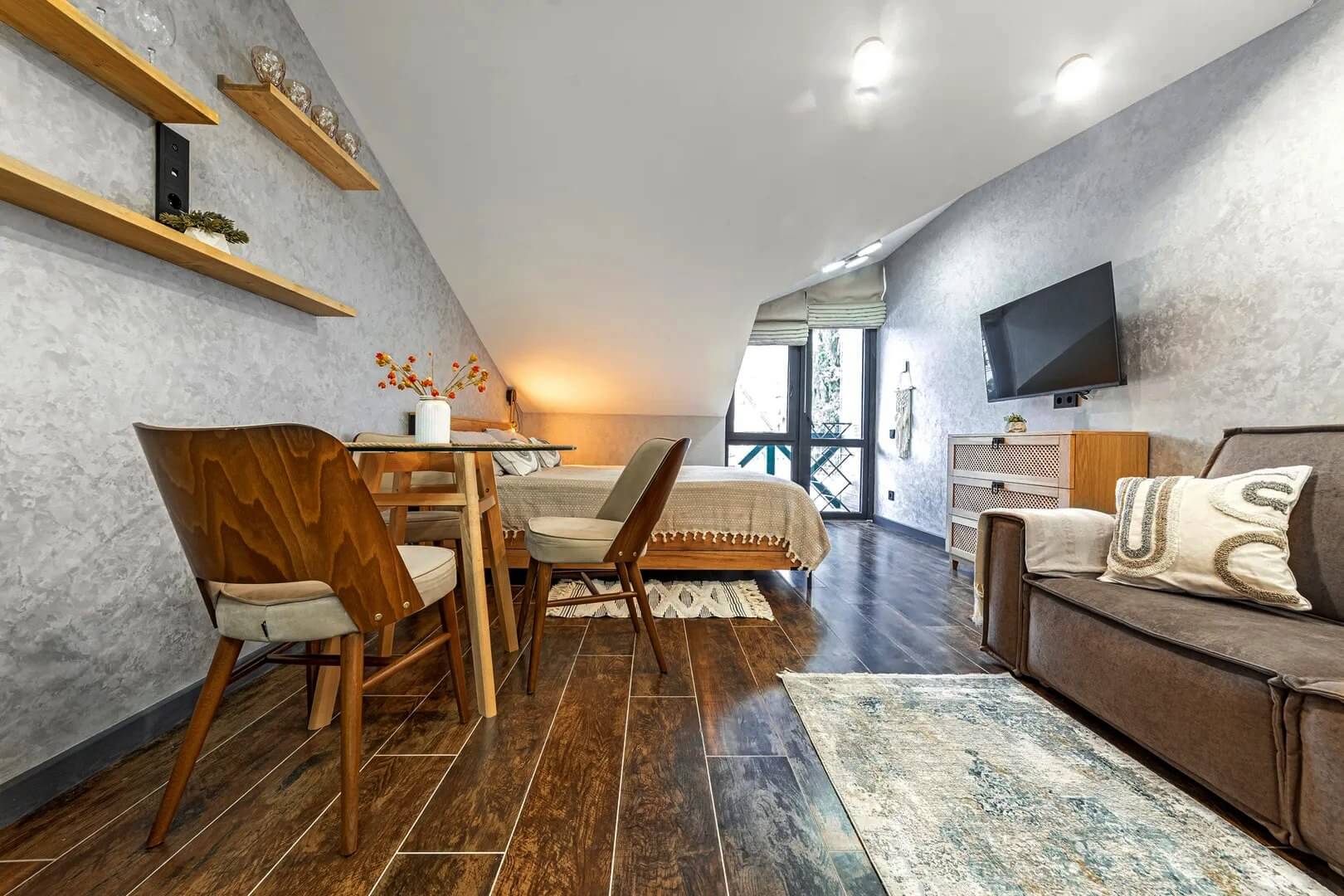 Панорама комнаты: диван с кухонным столом, кровать и телевизор на стене.
