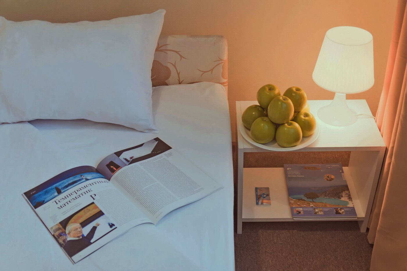 На кровати - журнал, на столе возле лампы - яблоки.