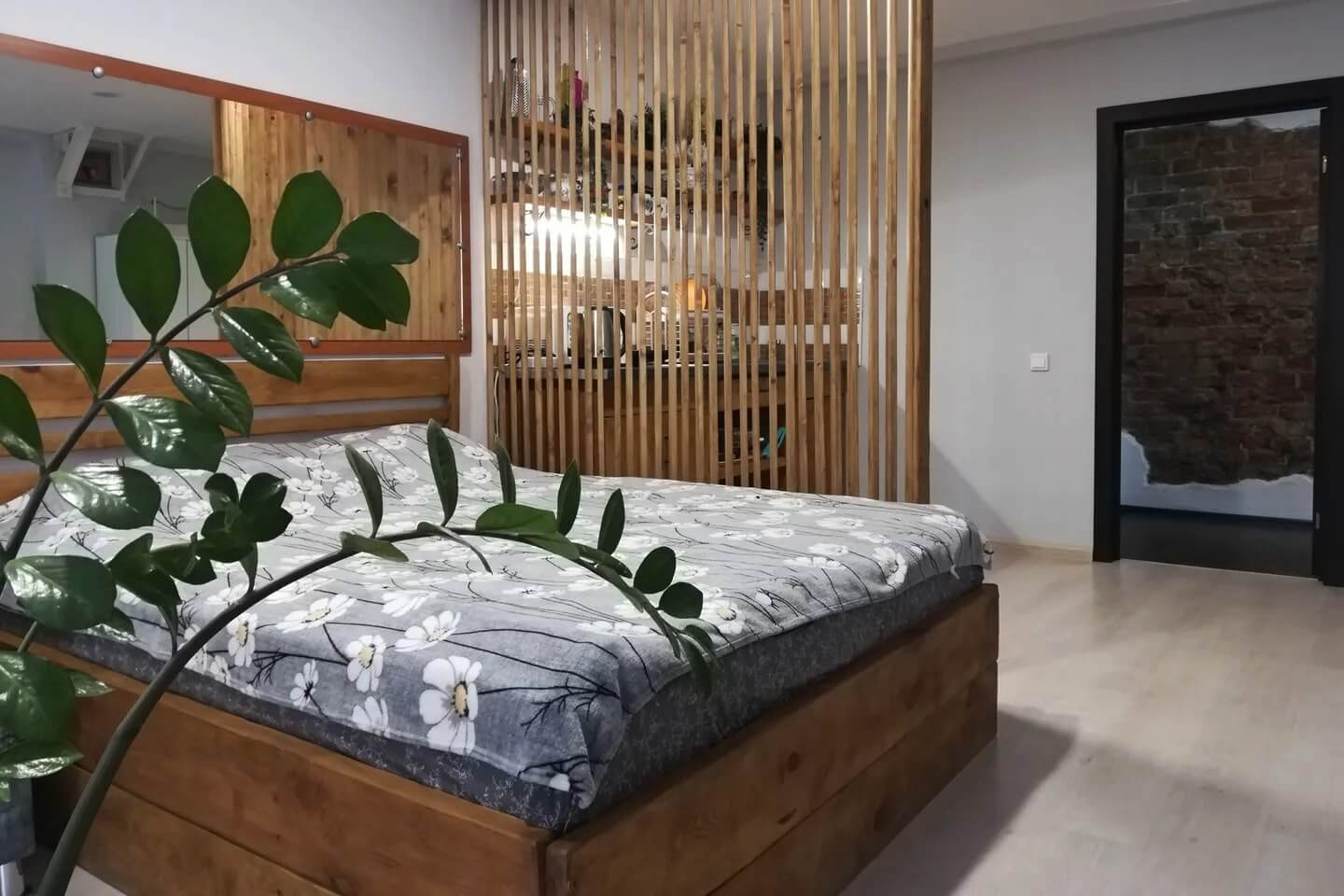 Спальное место: кровать, комнатное растение и деревянная перегородка.