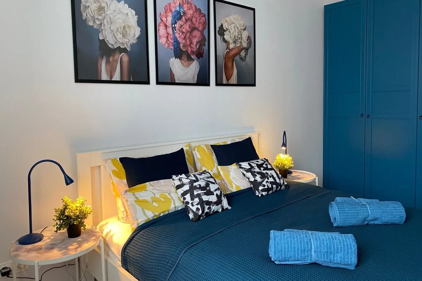 Спальное место: кровать с подушками, картины на стене, шкаф и покрывало "морского" синего цвета.