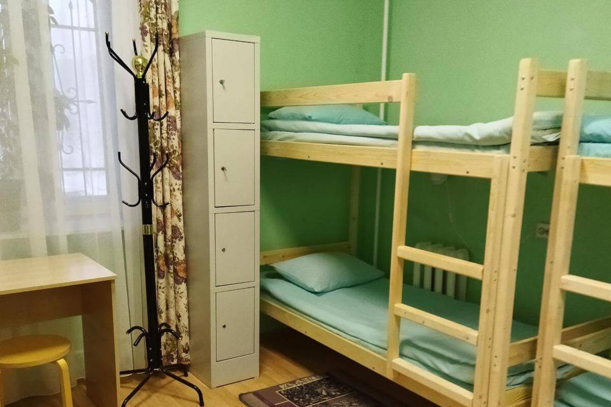 В комнате: небольшой столик, напольная вешалка, металлический шкафчик для личных вещей и деревянные двухъярусные кровати.