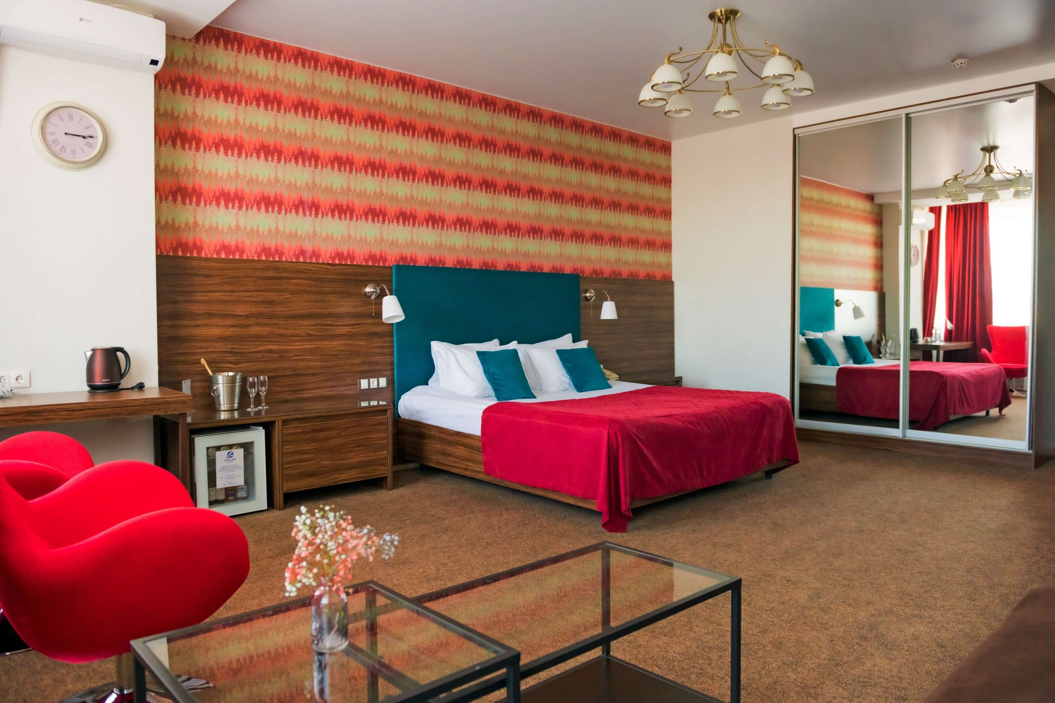 Гостиничный номер: большая кровать, шкаф-купе с зеркалом и два ярких красных кресла.