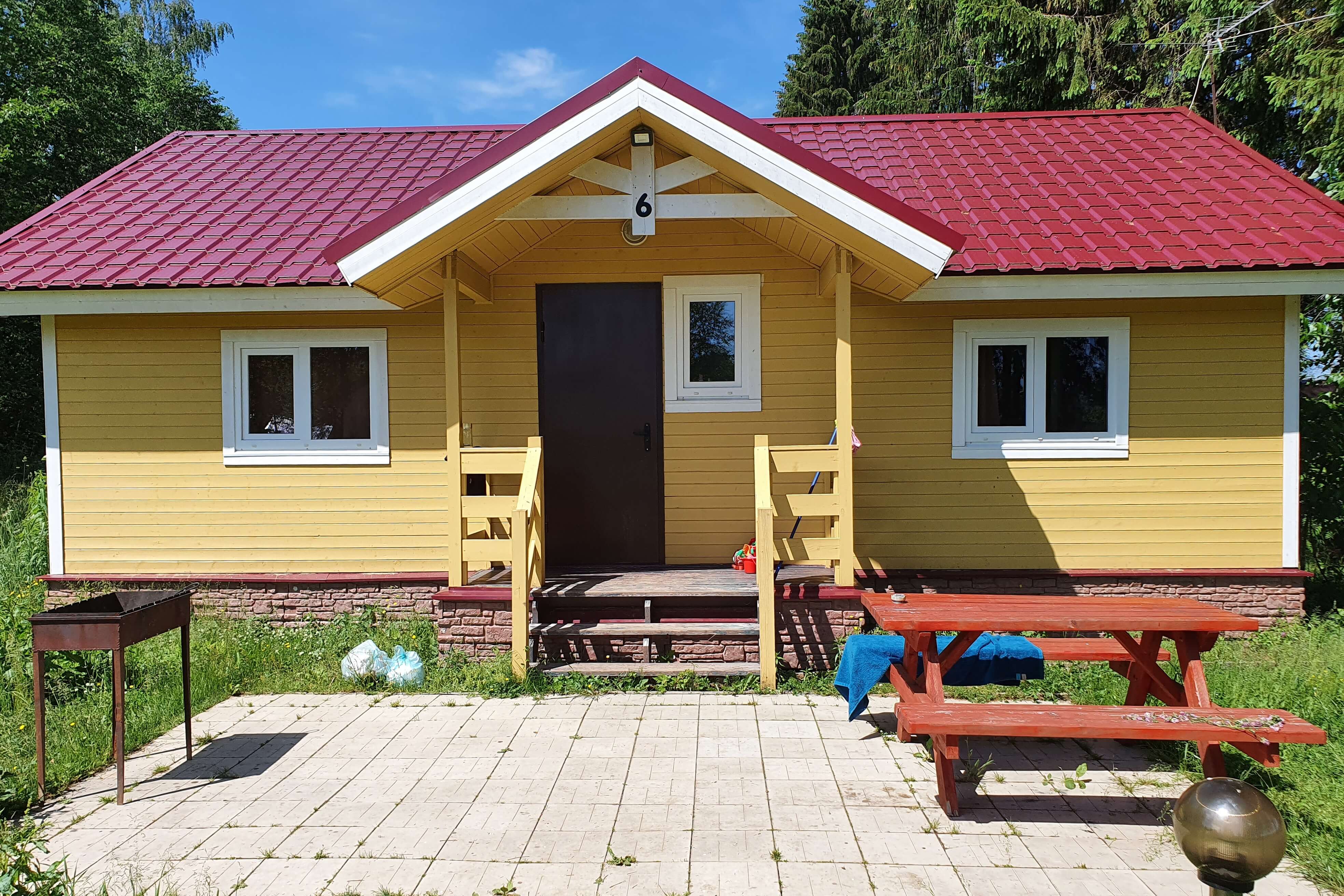 Фасад домика выкрашен в желтый цвет, перед крыльцом - мангал и столик для отдыха.
